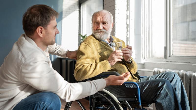 An old man is sitting in wheelchair near window and holding a glass of water. Next to him is a nurse.